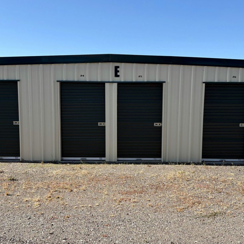 Beige metal storage building with dark roll-up doors on a gravel lot under a clear blue sky